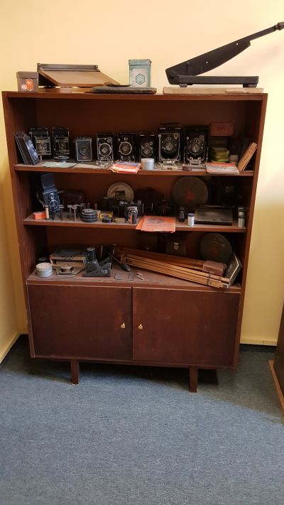 Dwie gilotyny do cięcia zdjęć (widoczne na g&oacute;rnej p&oacute;łce szafy), kolekcja aparat&oacute;w fotograficznych możliwych do kupienia w sklepie (pierwsze dwa aparaty od prawej strony na g&oacute;rnej p&oacute;łce to prywatne aparaty kliszowe Michała Franciszka Gottmana) oraz elementy diaskopu, drewniane statywy i klisze.  Zdjęcie udostępnione przez Pana Grzegorza Dąbrowskiego